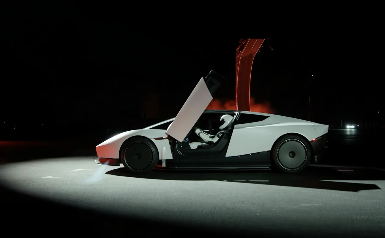 A white Tesla Roadster at night with its gull-wing driver door raised and a driver in a white helmet and suit inside, red light glowing behind